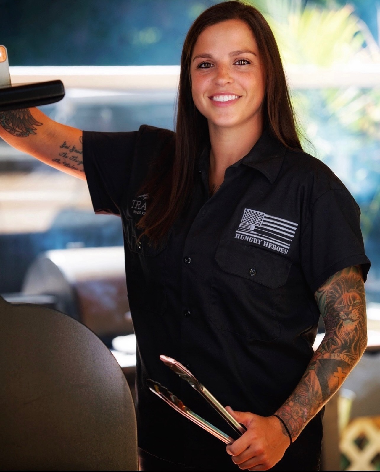 Girl standing holding grill tongs standing in front of an open grill wearing a black short-sleeved button down shirt with long brown hair and smiling.